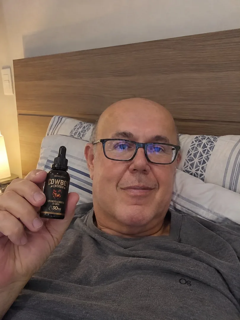 Homem de óculos deitado na cama, segurando um frasco COWBOY Energia