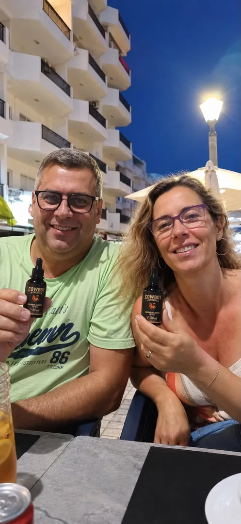 Casal sorrindo em uma mesa ao ar livre à noite, cada um segurando um frasco COWBOY Energia